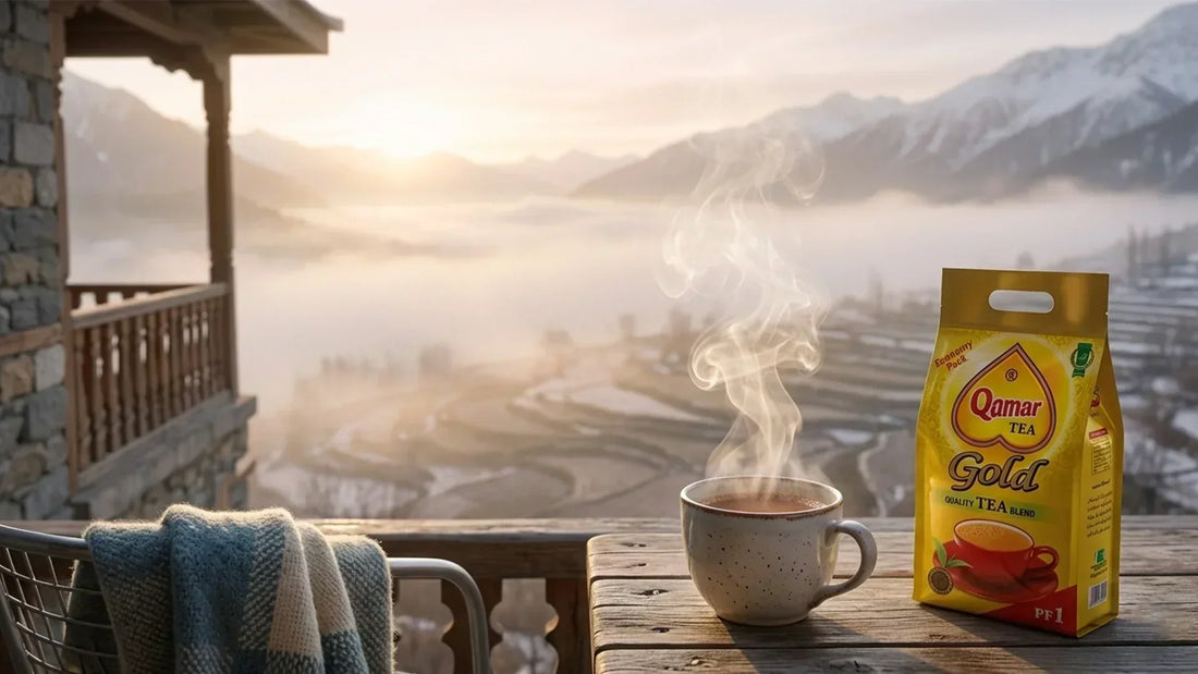 A steaming mug of tea next to a yellow pack of Qamar Gold tea on a balcony overlooking misty mountains.